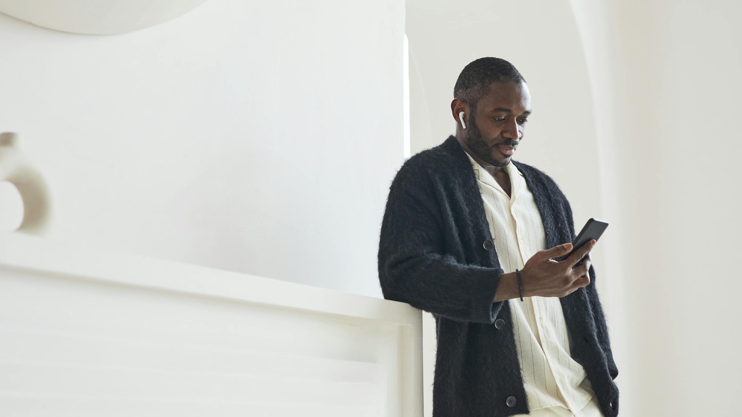 Fashionably dressed man using smartphone and earbuds indoors.
