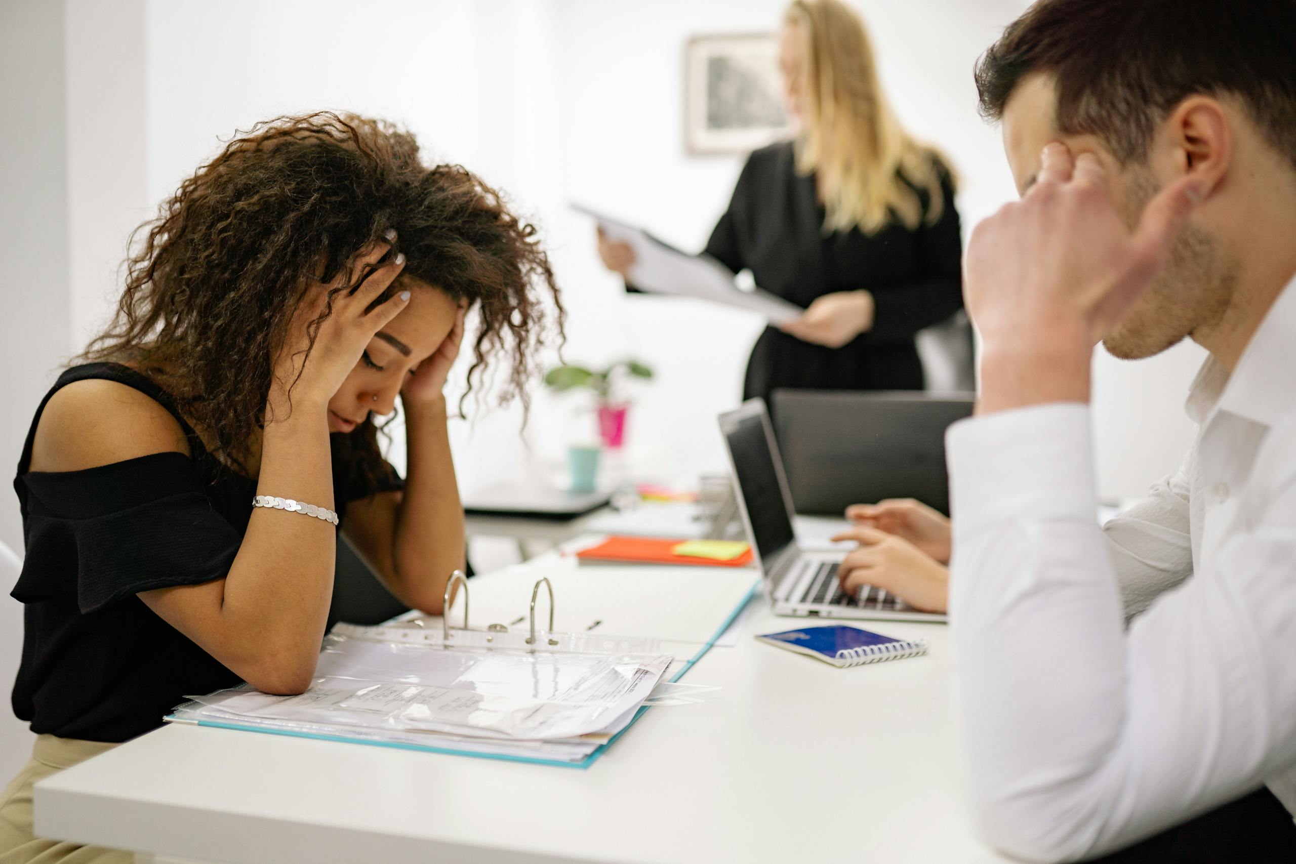 African American woman stressed at office desk with nearby colleagues conveying workplace pressure