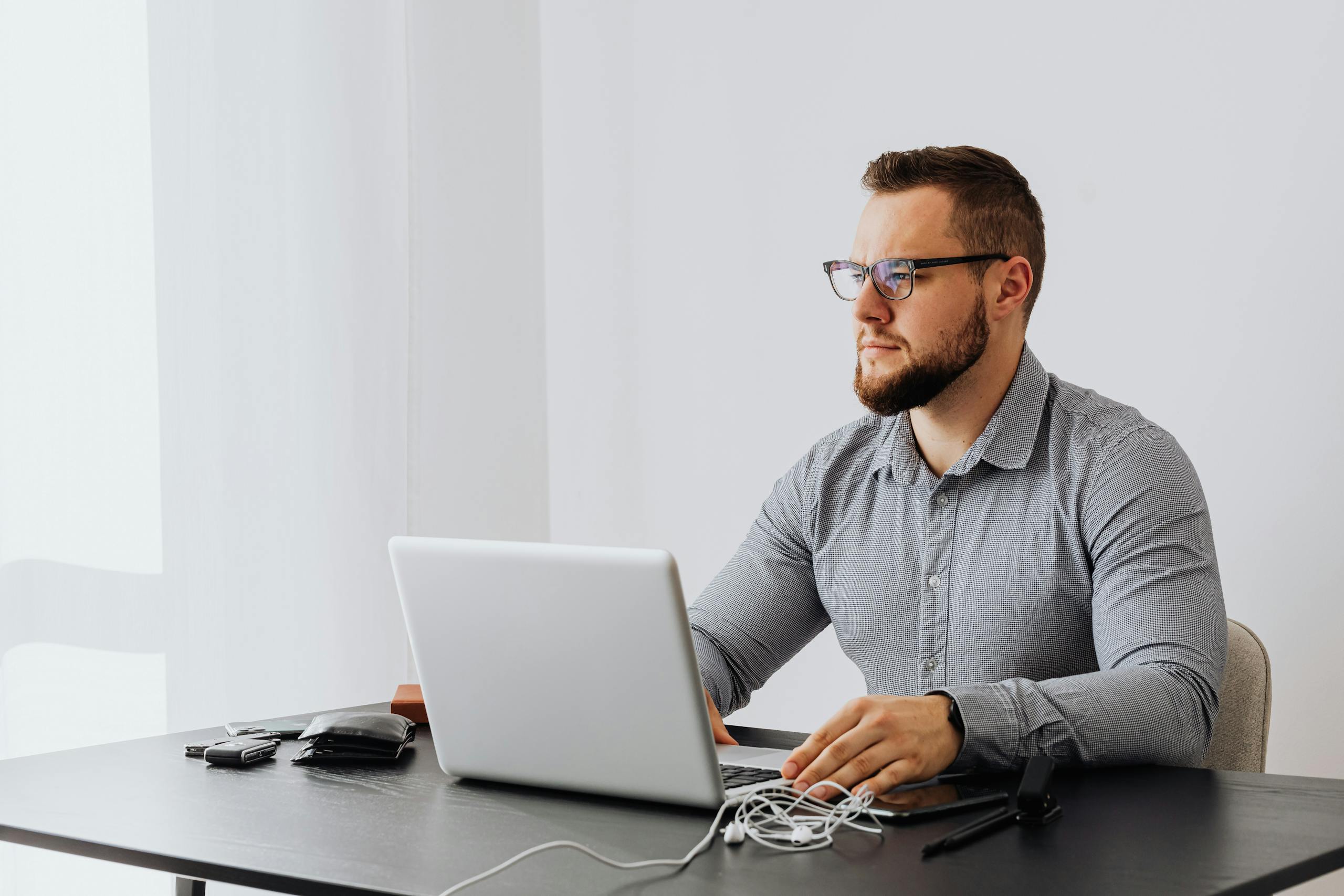 Bearded man with eyeglasses working on laptop in minimalist office setting.