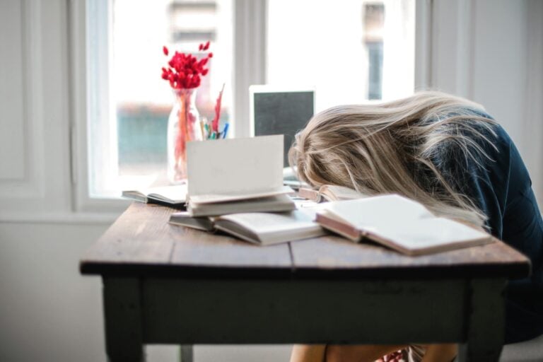 Blonde woman slumped over desk surrounded by books showing study fatigue.