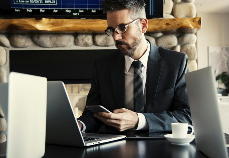 Businessman in formal attire working on laptop and smartphone at office desk.