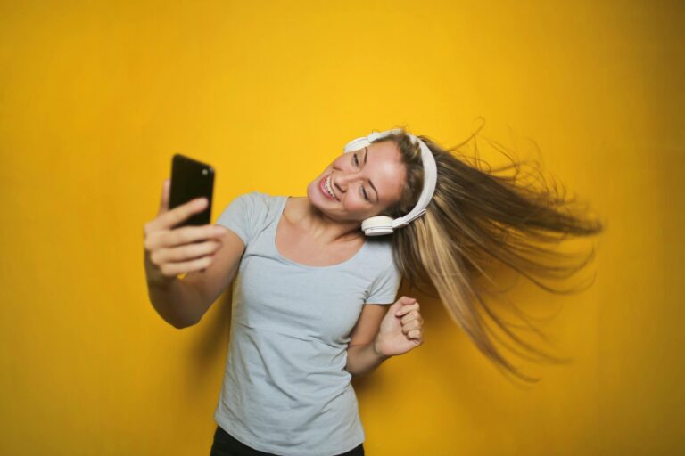 Cheerful woman with headphones enjoying music and taking selfie against bright yellow background.