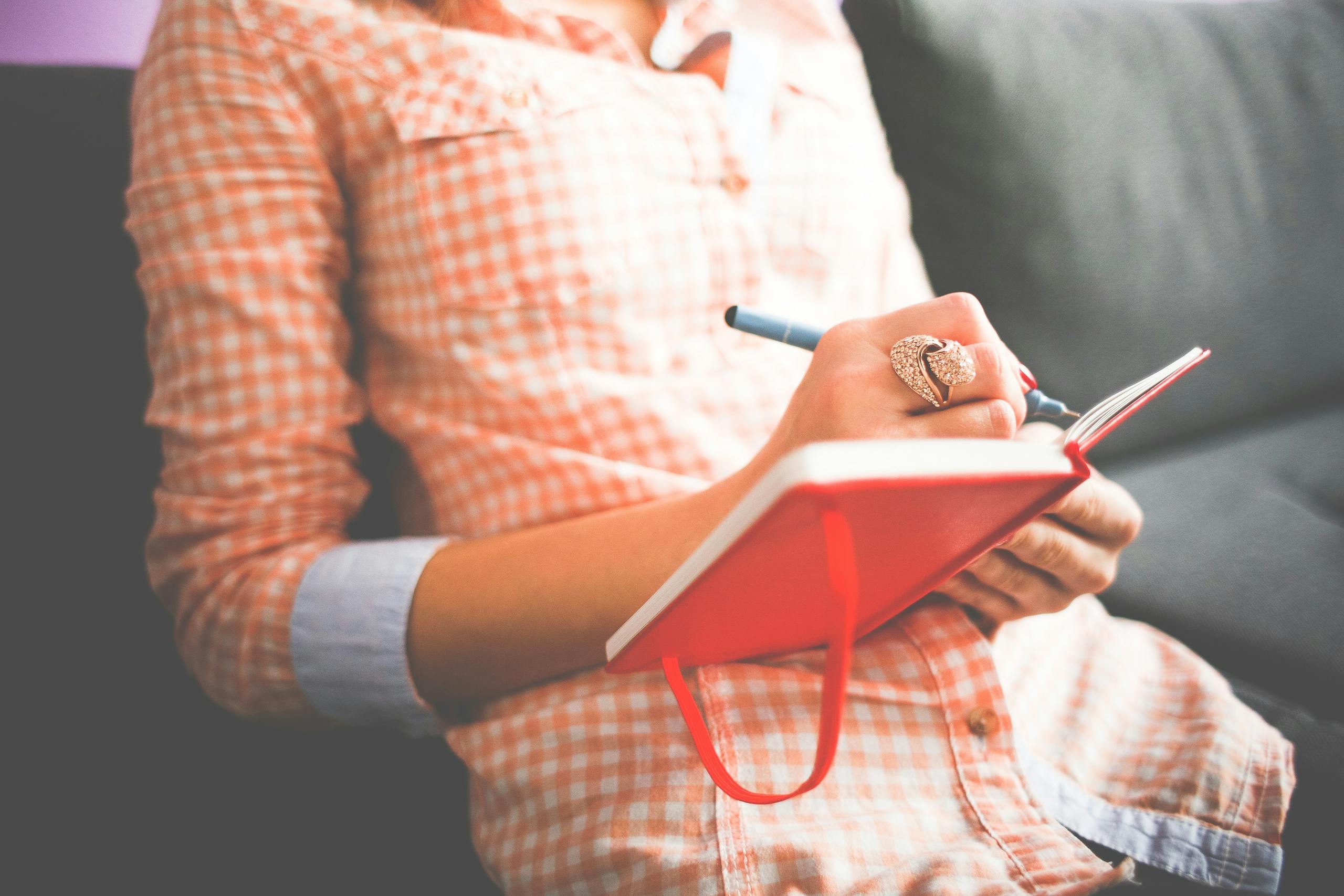 Woman writing in red journal on sofa wearing stylish ring