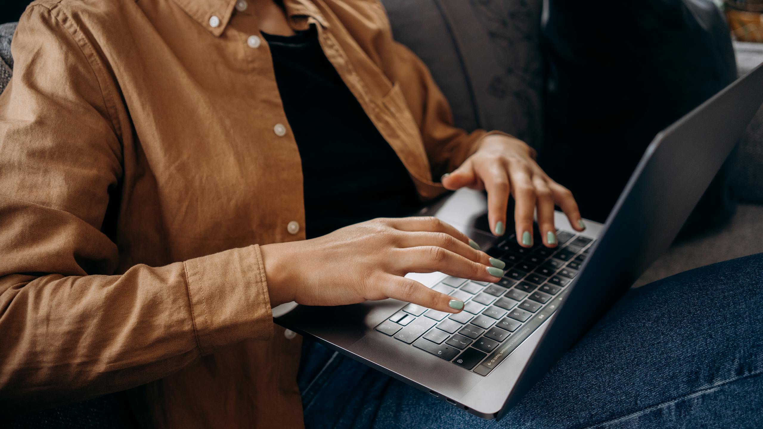 Close-up of hands typing on laptop keyboard while wearing brown casual shirt