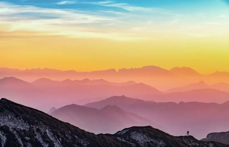 Colorful sunrise over misty mountains with lone hiker silhouette visible.