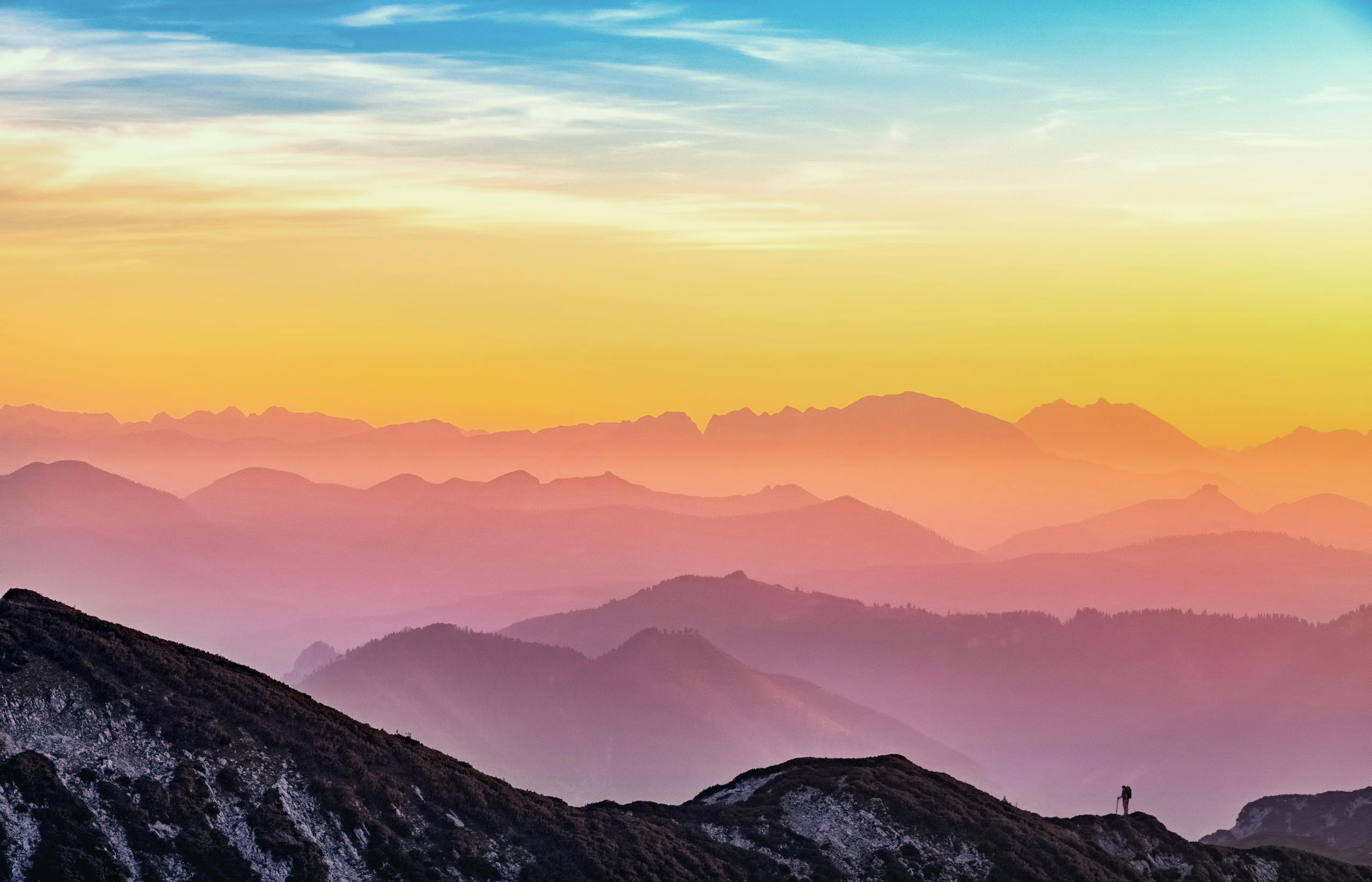 Colorful sunrise over misty mountains with lone hiker silhouette visible.