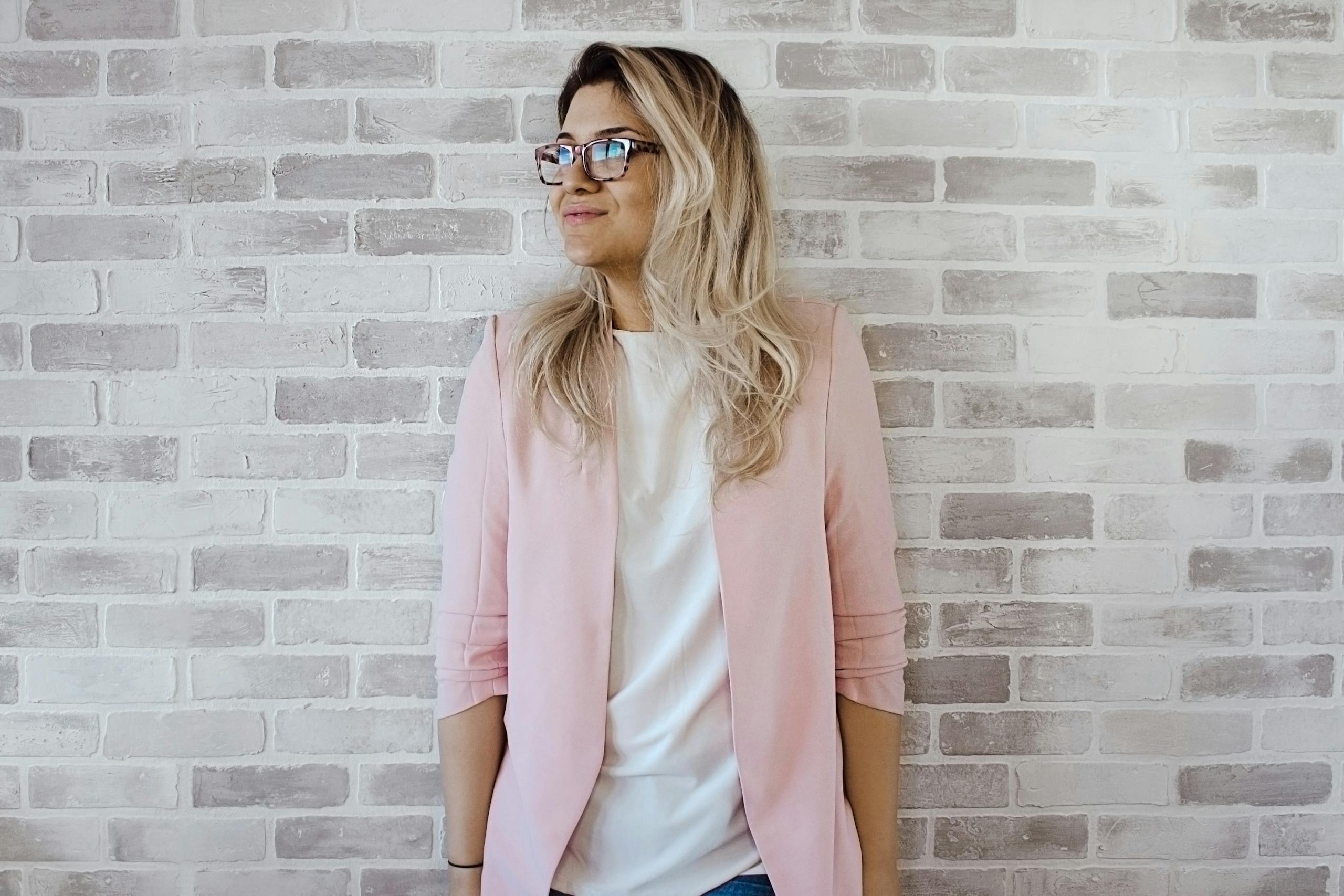 Elegant woman wearing glasses and pink blazer posing against brick wall backdrop.