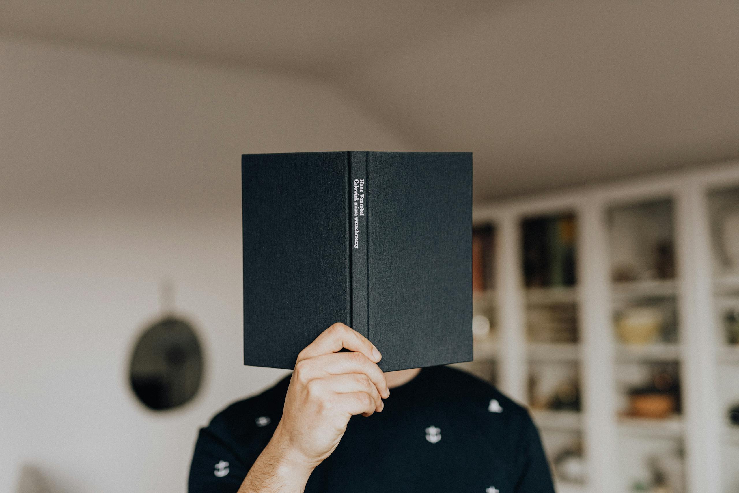 Faceless man holding open black book covering face in bright room