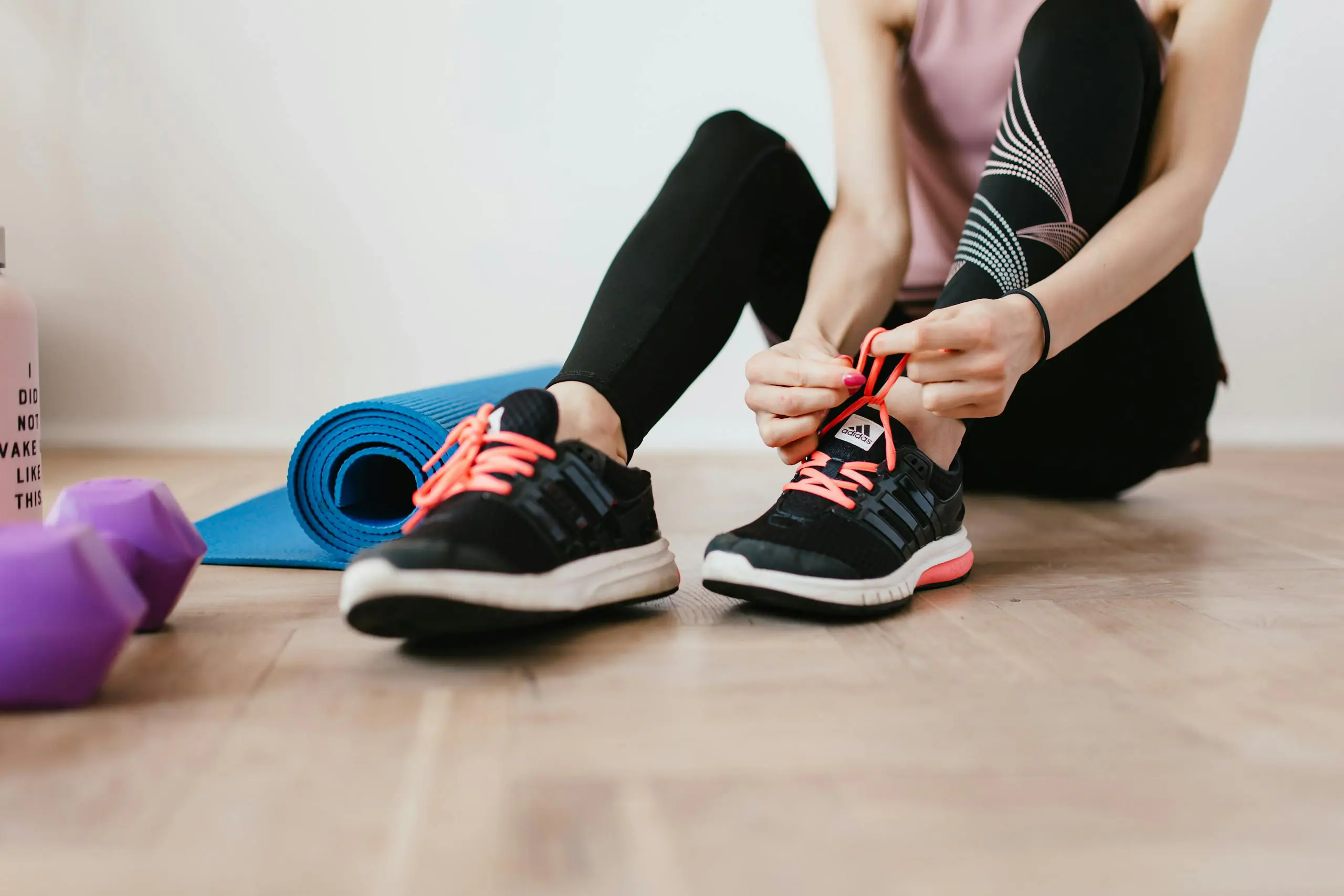 Woman tying shoelaces before indoor workout with yoga mat and dumbbells.