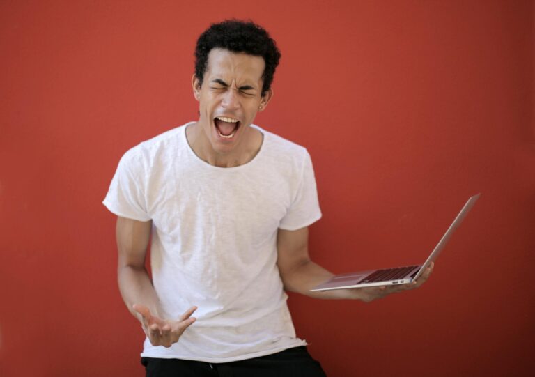 Man with open mouth and closed eyes screaming madly with laptop on red background.