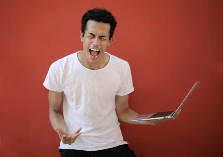 Man with open mouth and closed eyes screaming madly with laptop on red background.