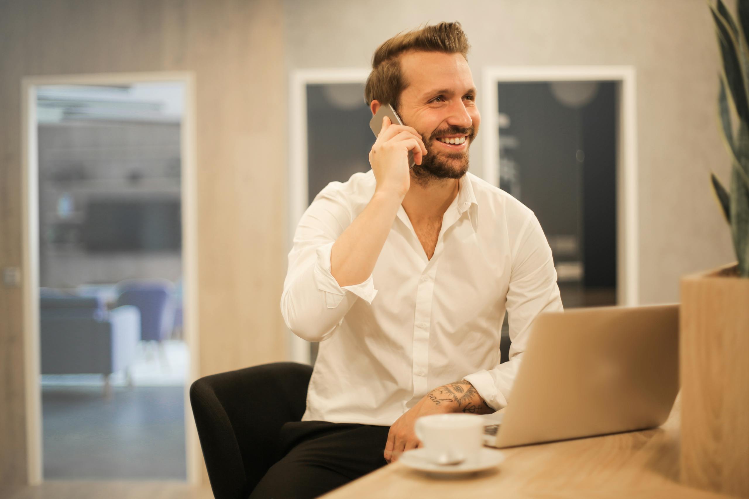 Happy businessman in white shirt on phone call at modern office desk with laptop