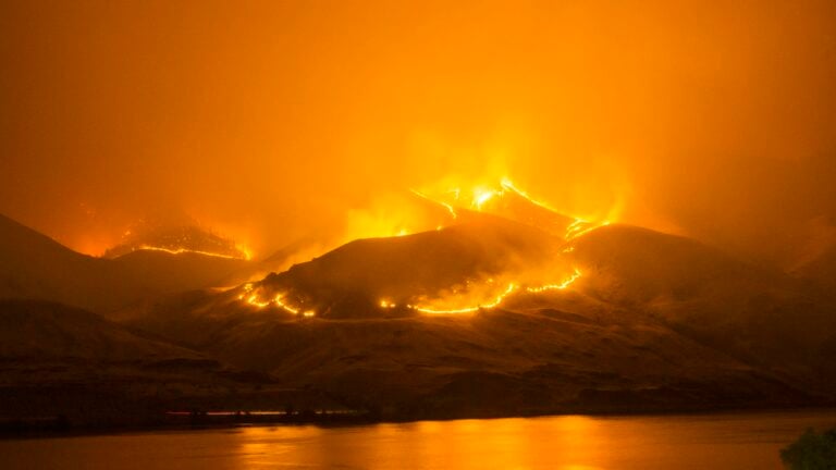 Intense wildfire blazing on hills reflected in calm river below.