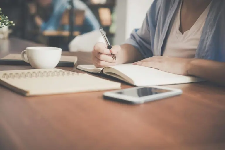Woman writing in notebook at desk with coffee cup and phone nearby