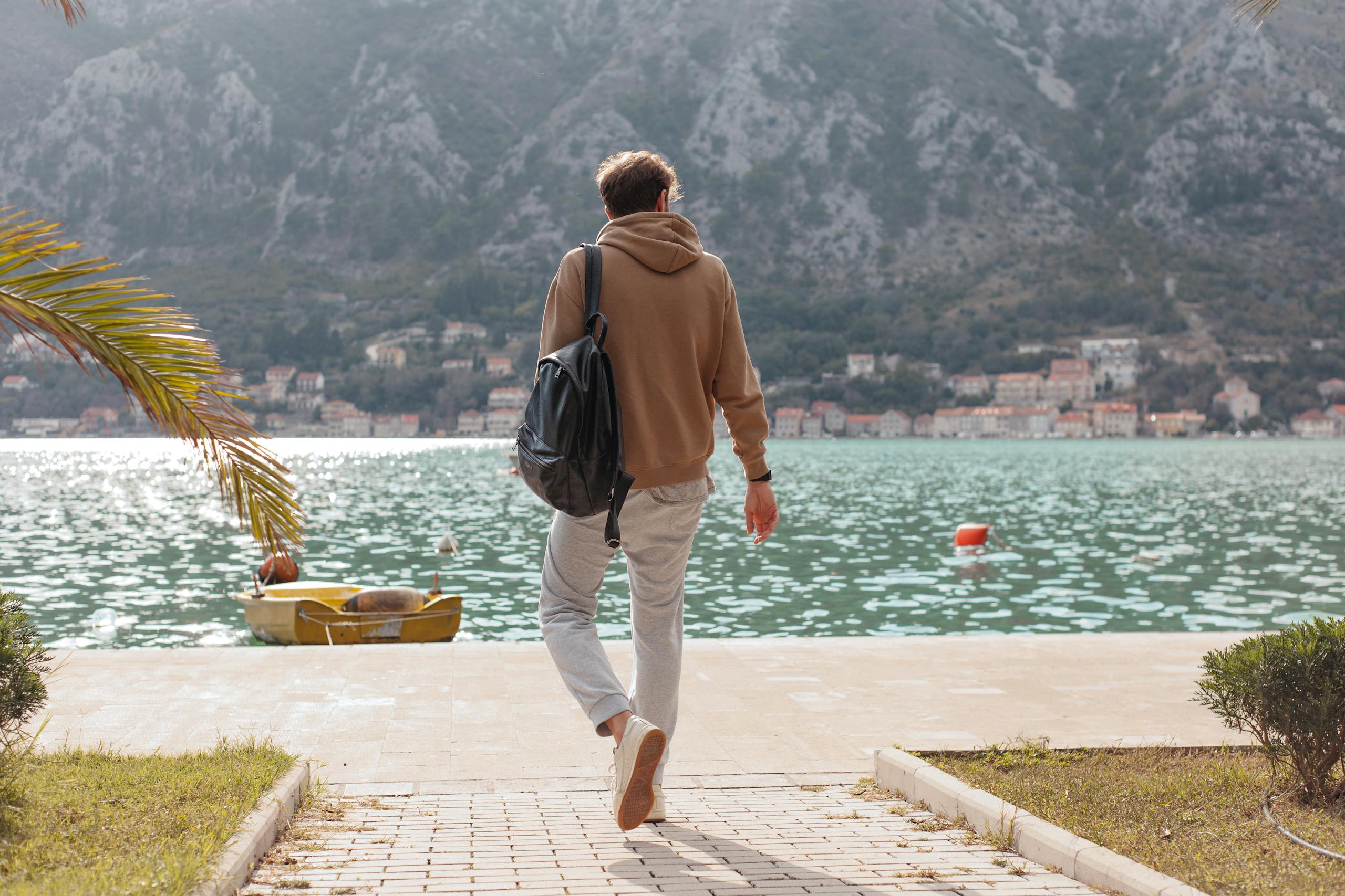 Man in hoodie walking on dock towards calm waterfront with mountains.