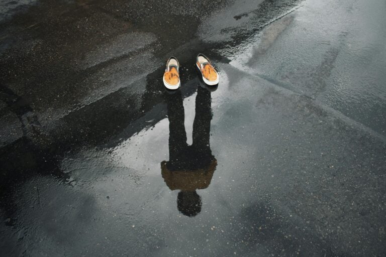 Reflection of legs and shoes in puddle on wet urban street creating surreal scene.