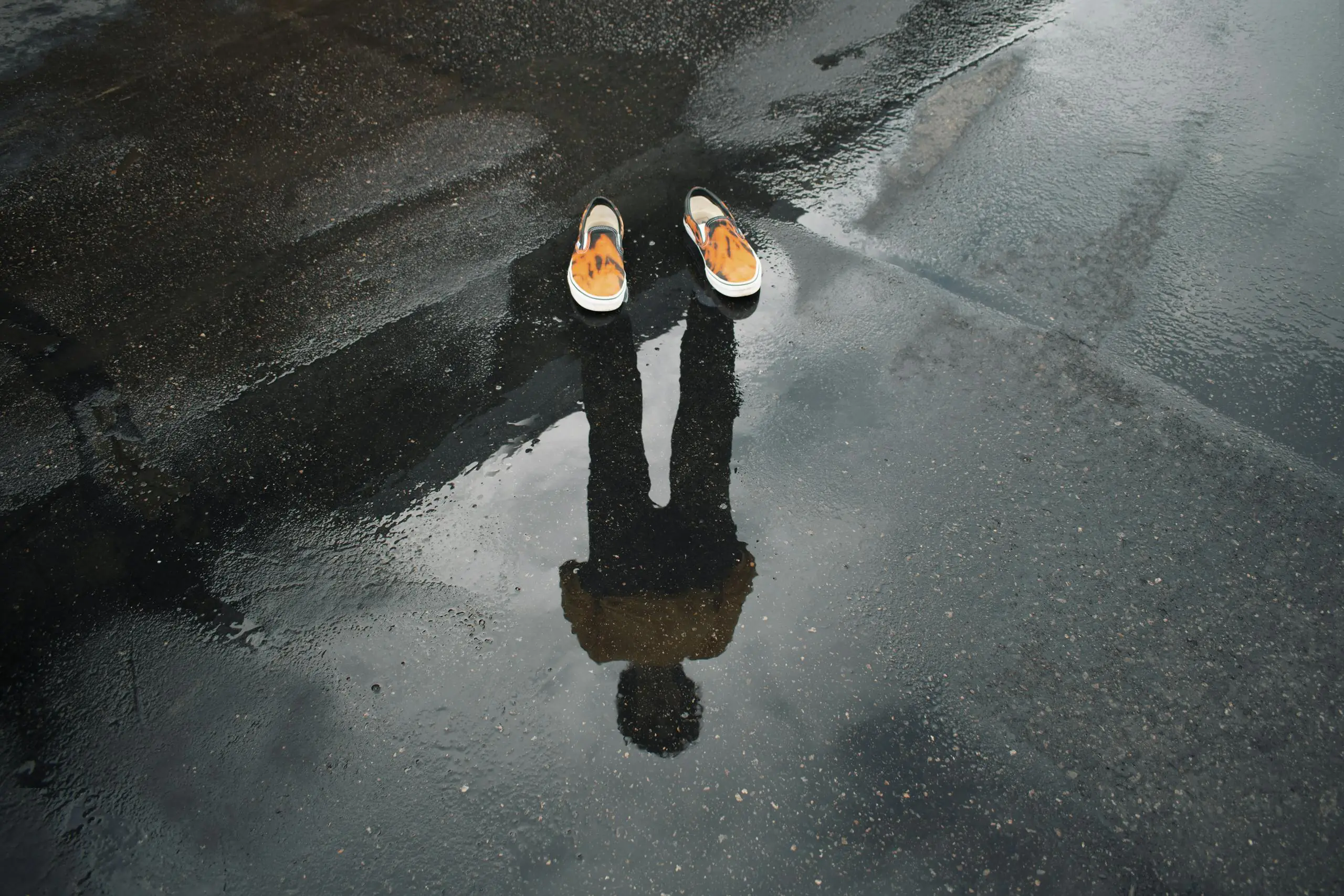 Reflection of legs and shoes in puddle on wet urban street creating surreal scene.