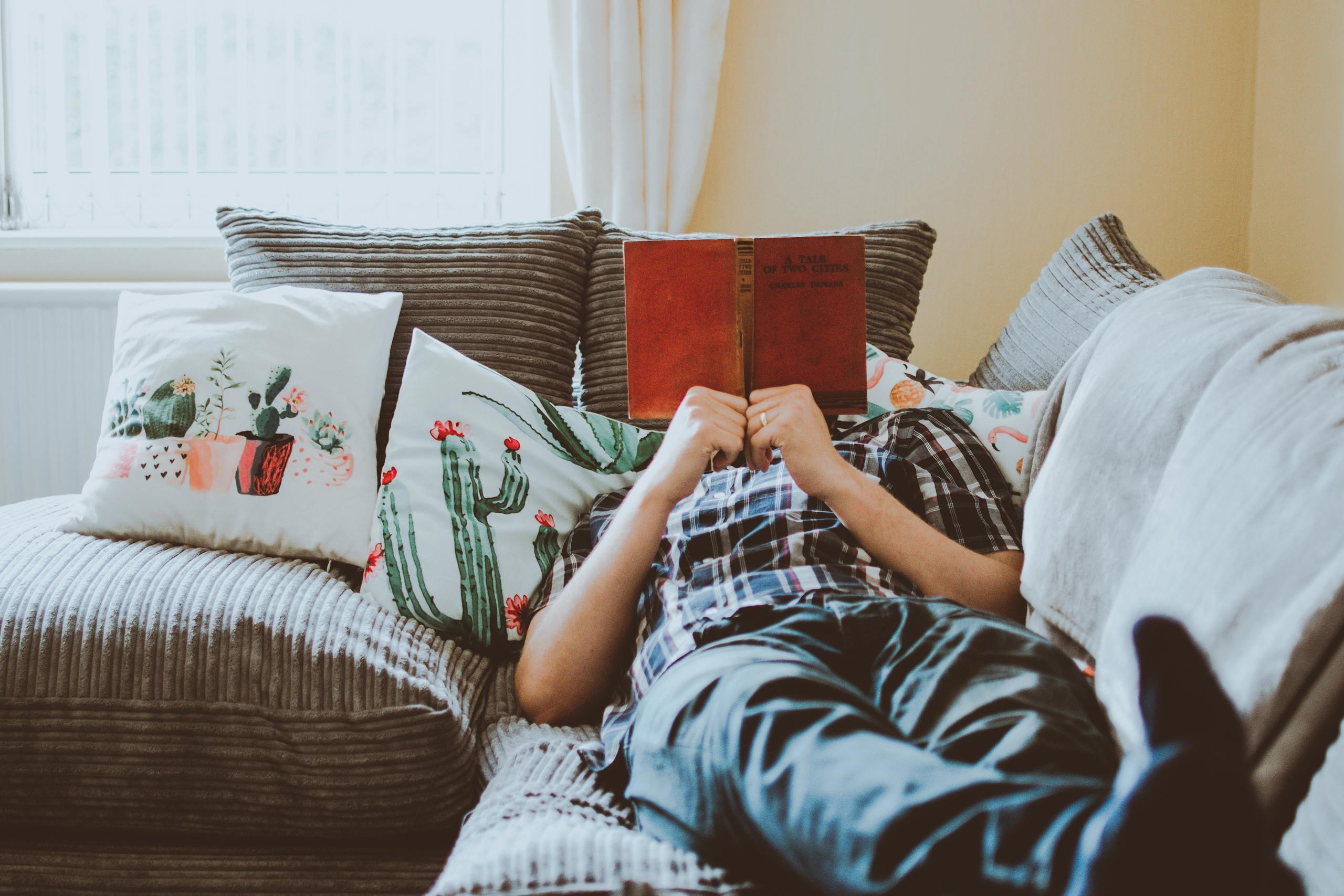 Relaxed person lying on sofa reading book surrounded by soft cushions.