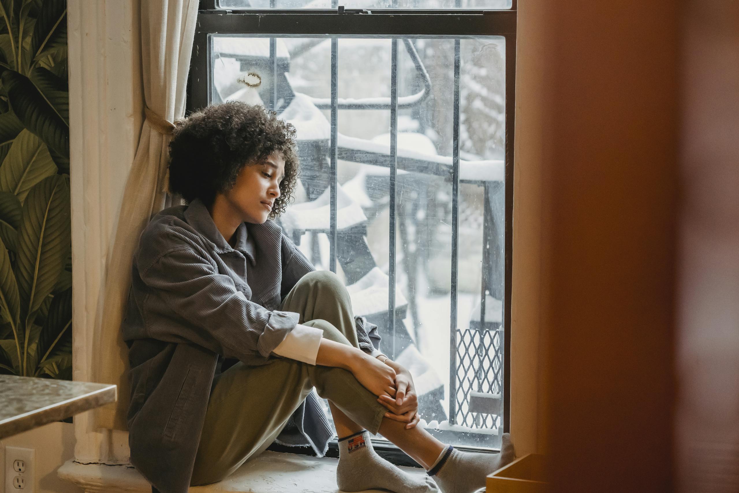 Upset African American woman sitting in solitude near window embracing knees looking down.
