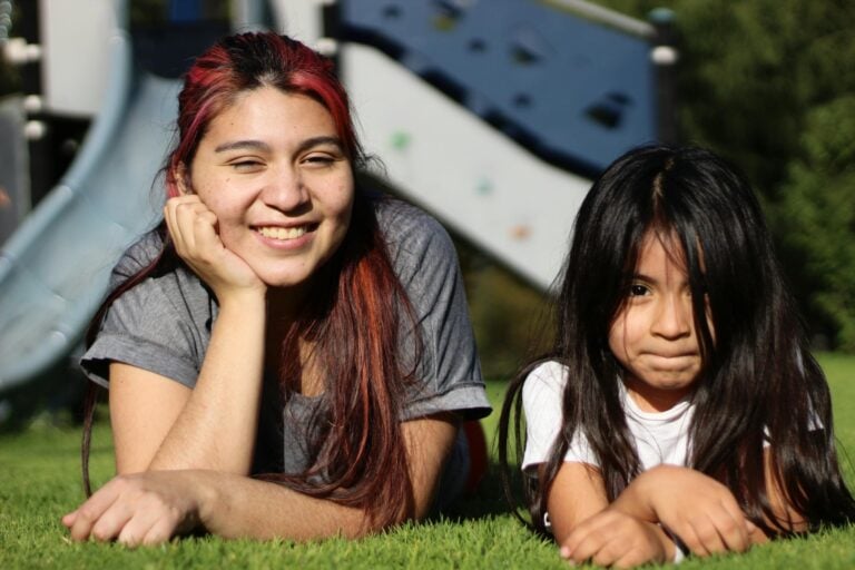Smiling mother and daughter relaxing on grass in sunny playground.