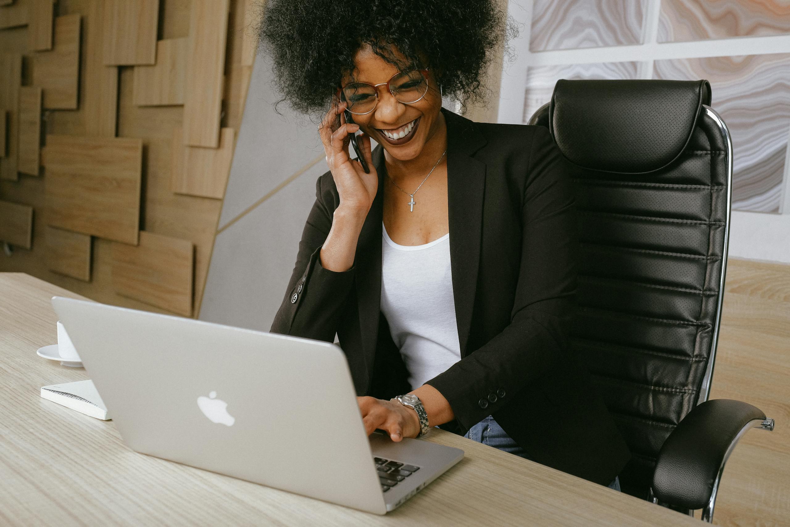 Smiling woman on phone call while working on laptop in stylish office.
