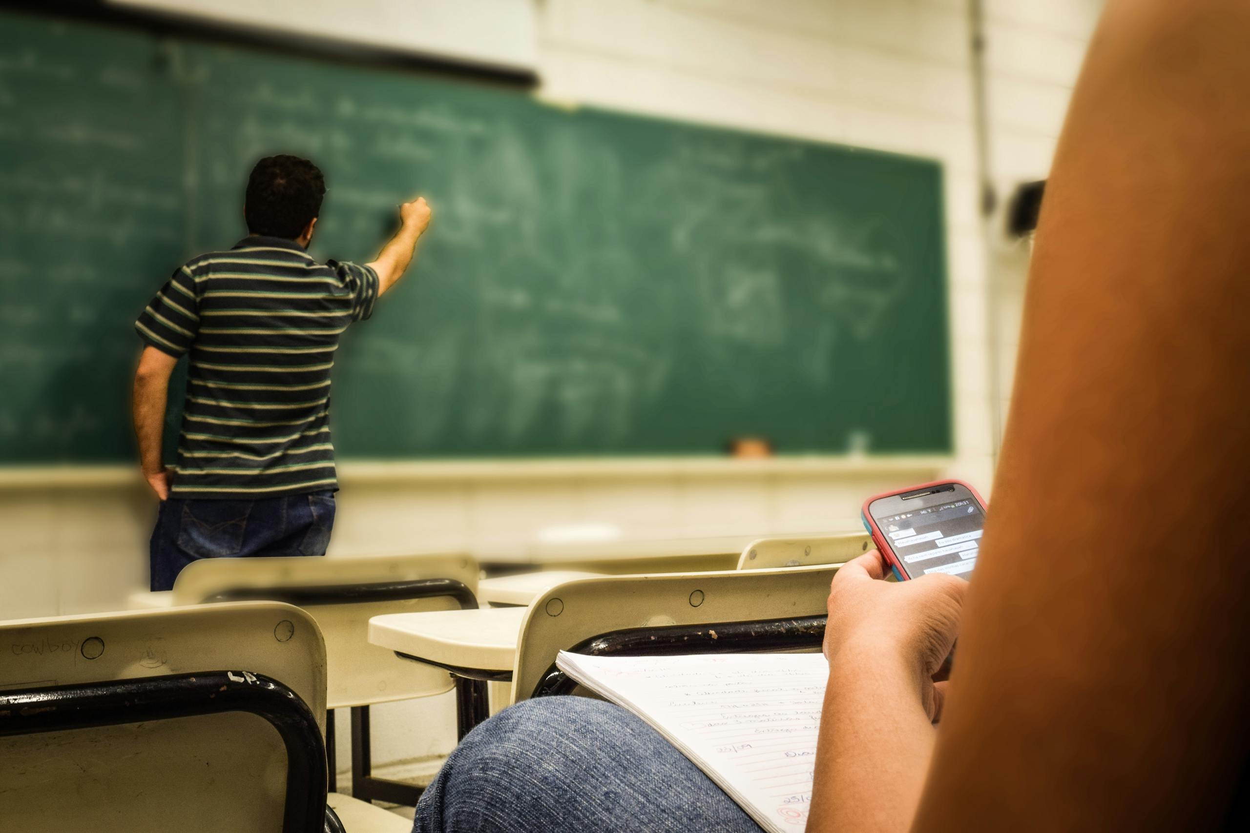 Student texting on phone in classroom while teacher writes on blackboard