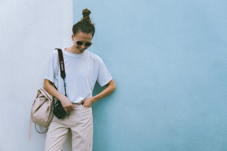 stylish woman posing with camera in casual attire against a blue wall.