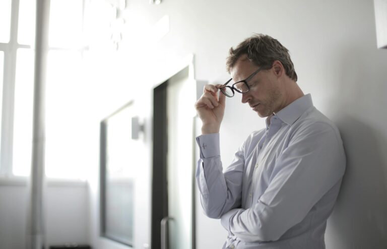 Thoughtful man in bright room holding glasses against wall contemplating