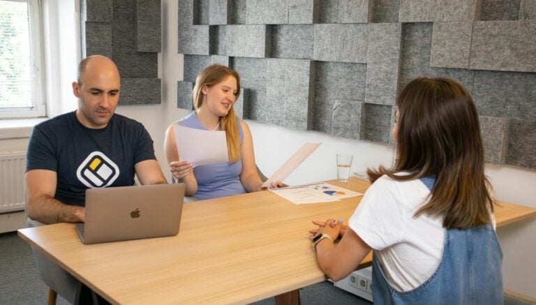 Three colleagues in modern office discussing work with laptops and documents present.