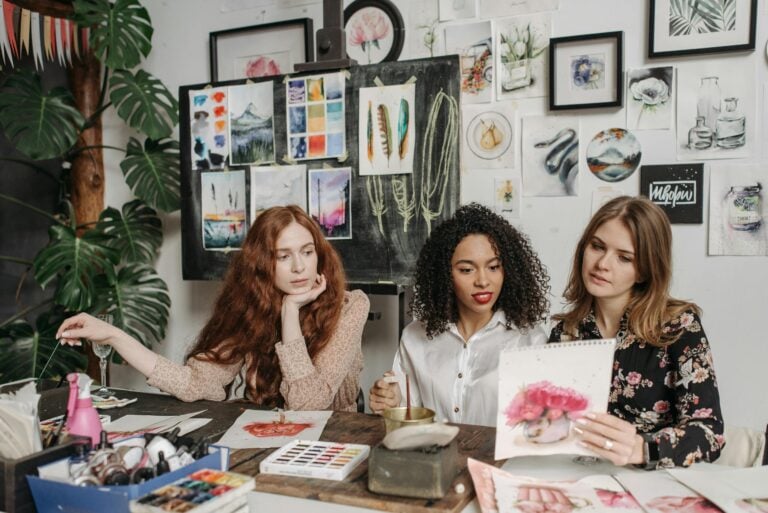 Three women discussing watercolor artwork in art studio surrounded by materials