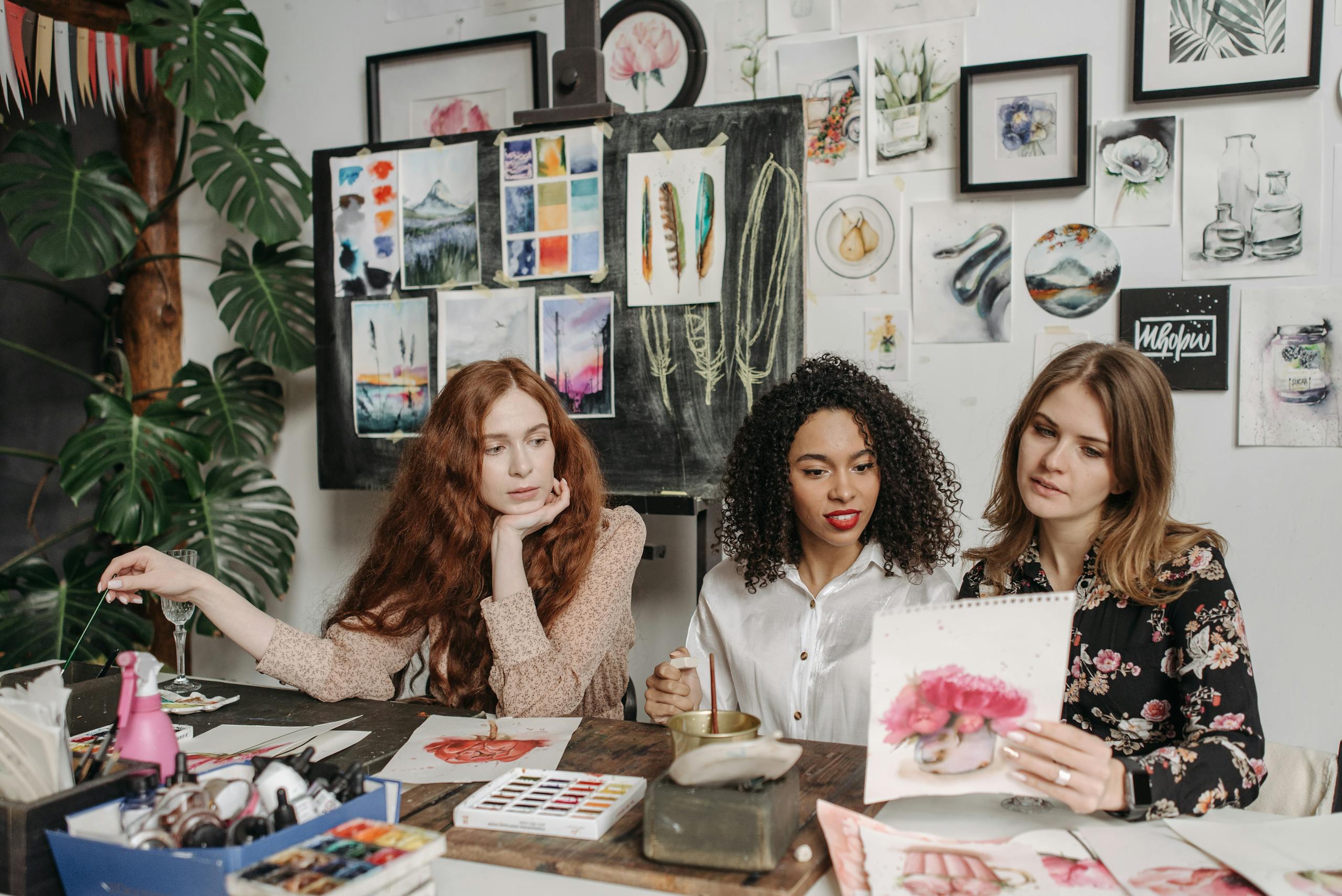 Three women discussing watercolor artwork in art studio surrounded by materials