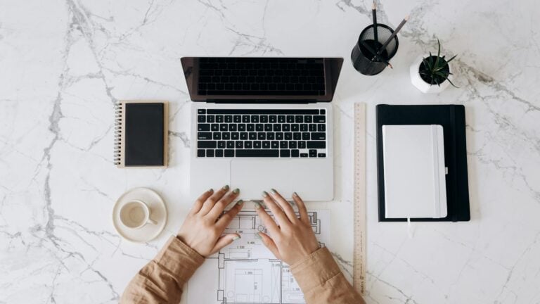 Top view of stylish home office desk with laptop, planner, coffee cup, and blueprint hands.