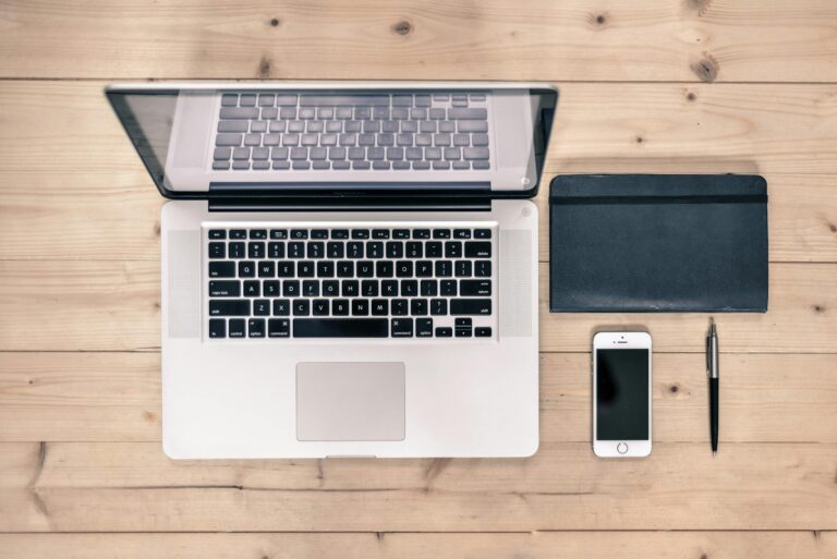 Top view of tidy workspace with laptop, smartphone, notebook, and pen on wooden desk.