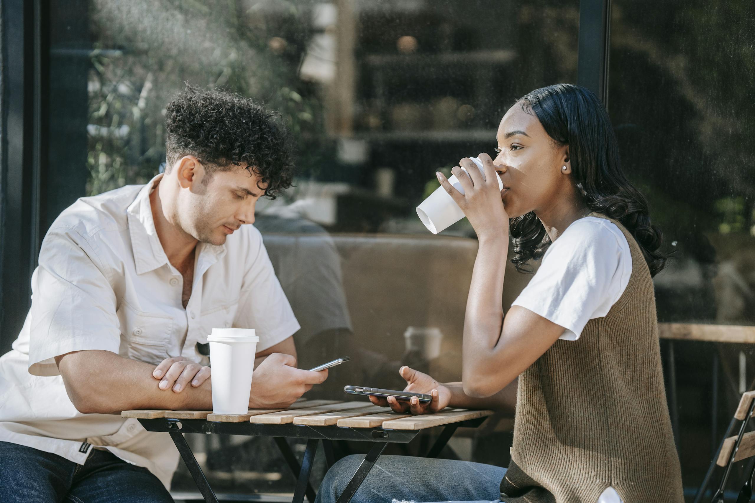 Two friends enjoying casual coffee break outdoors with smartphones in close-up view