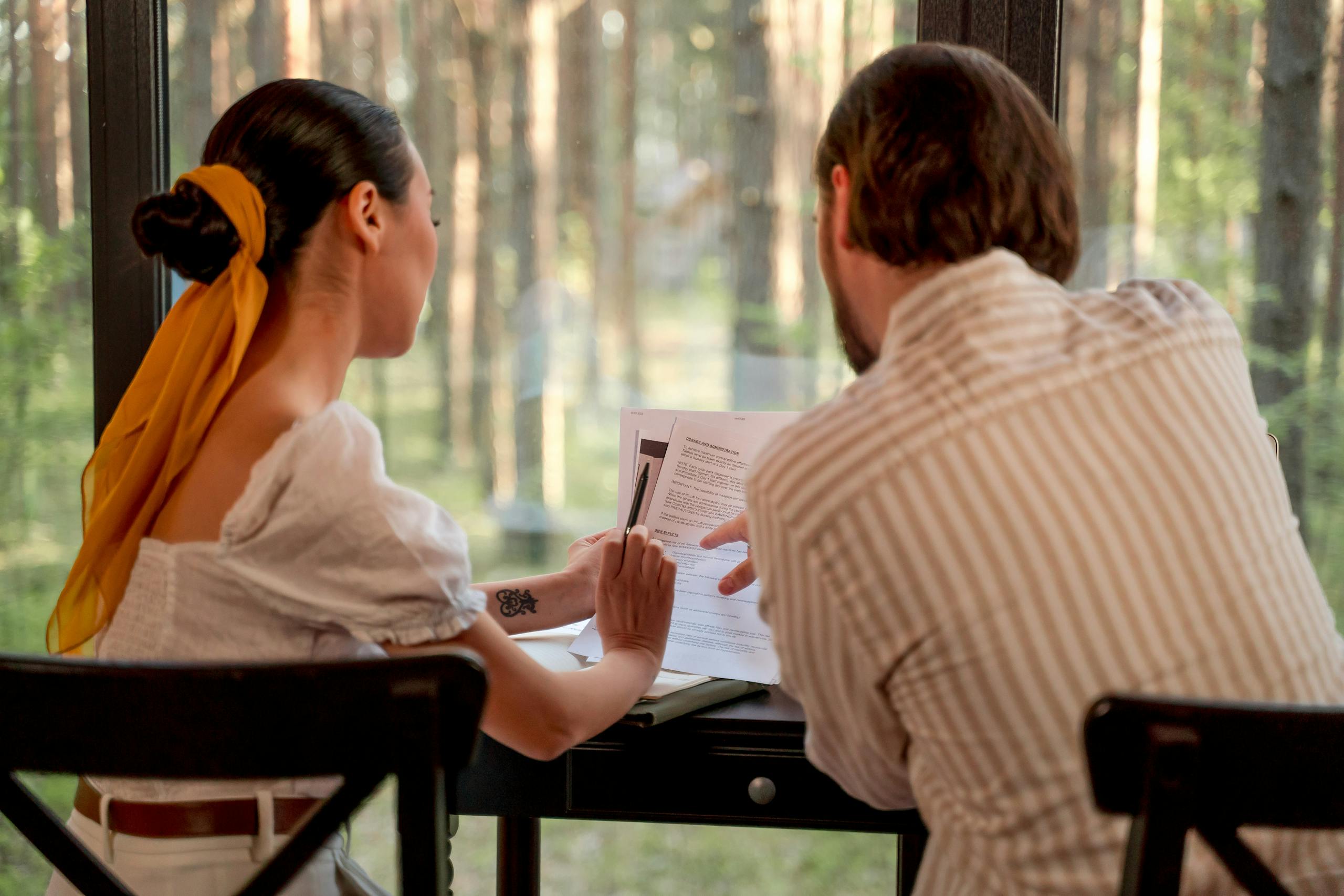 Two individuals collaborating on documents by large window with forest view.