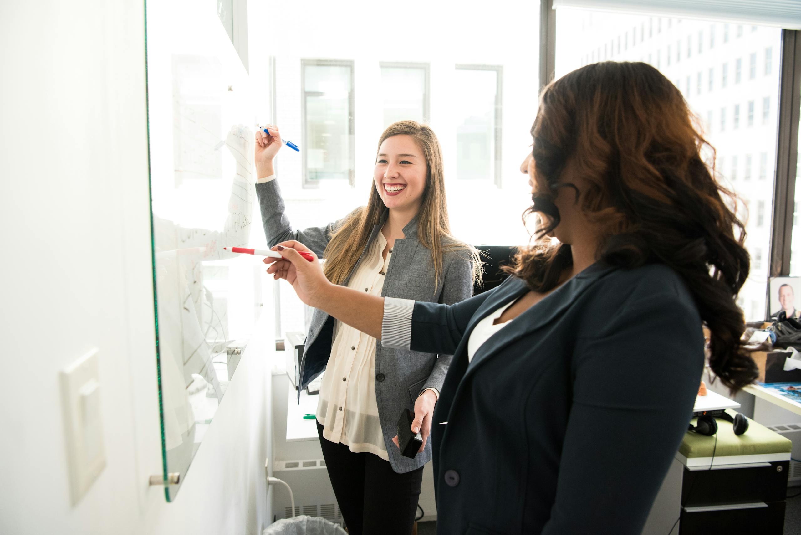 Two professional women discussing ideas on whiteboard in modern office setting.