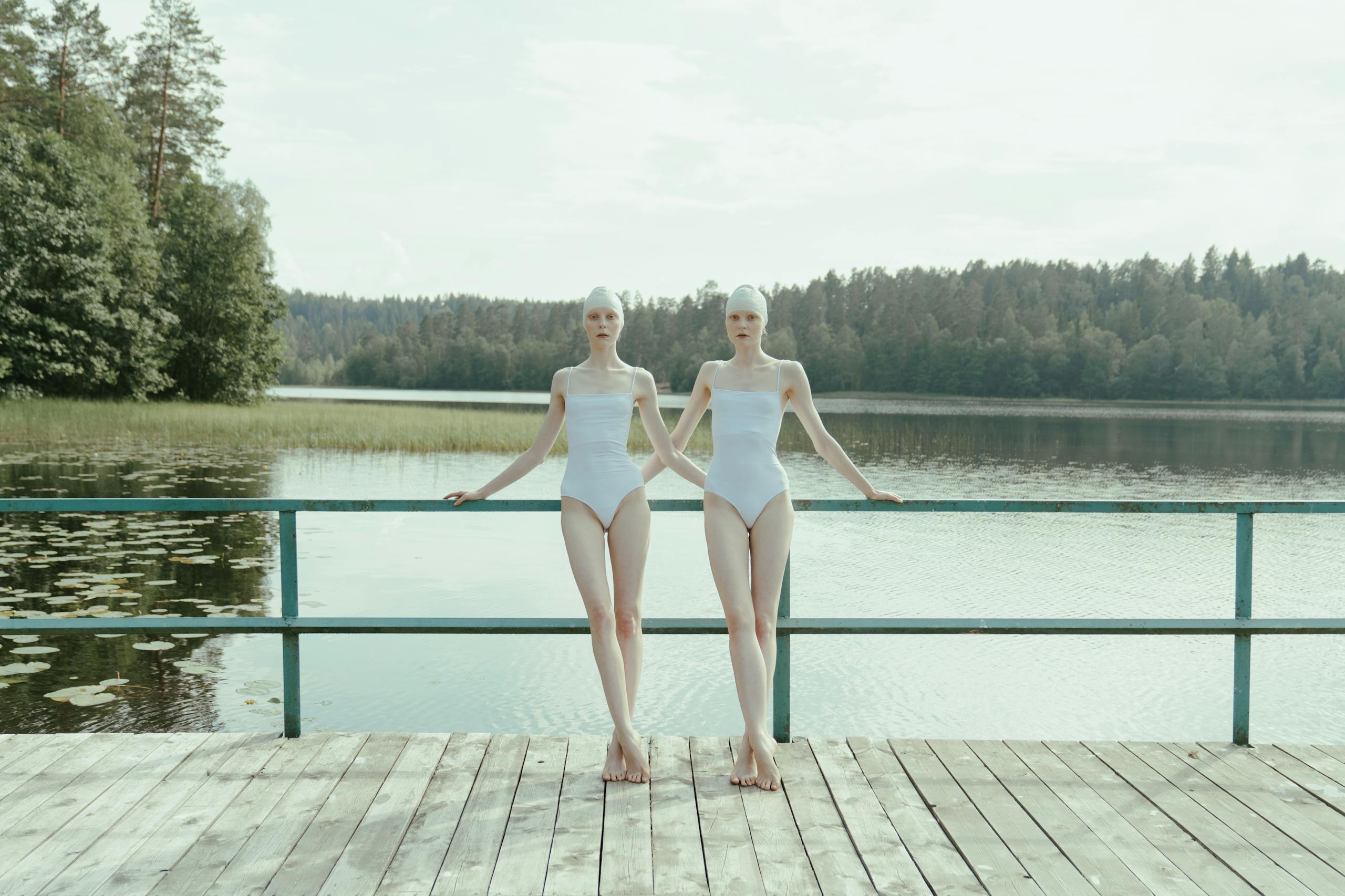 Two women in white swimsuits standing on dock at serene lake surrounded by trees.