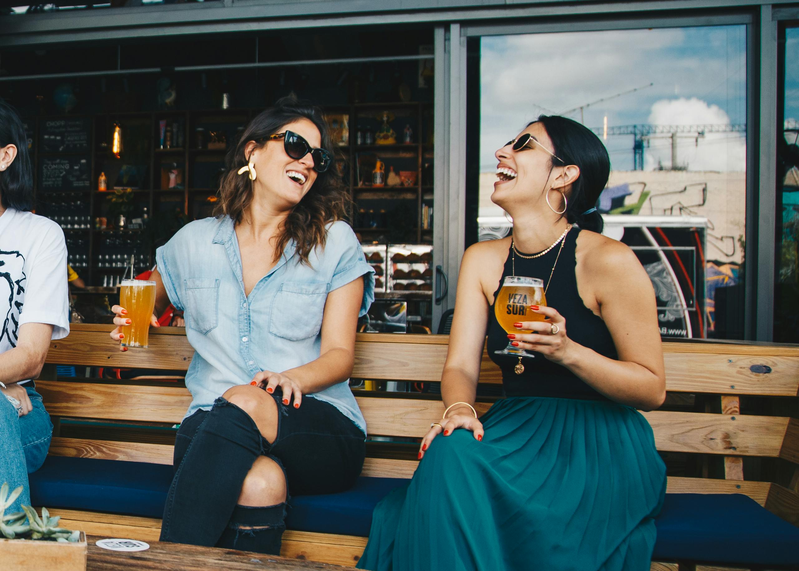 Two women laughing and enjoying drinks outdoors at trendy bar in vibrant setting