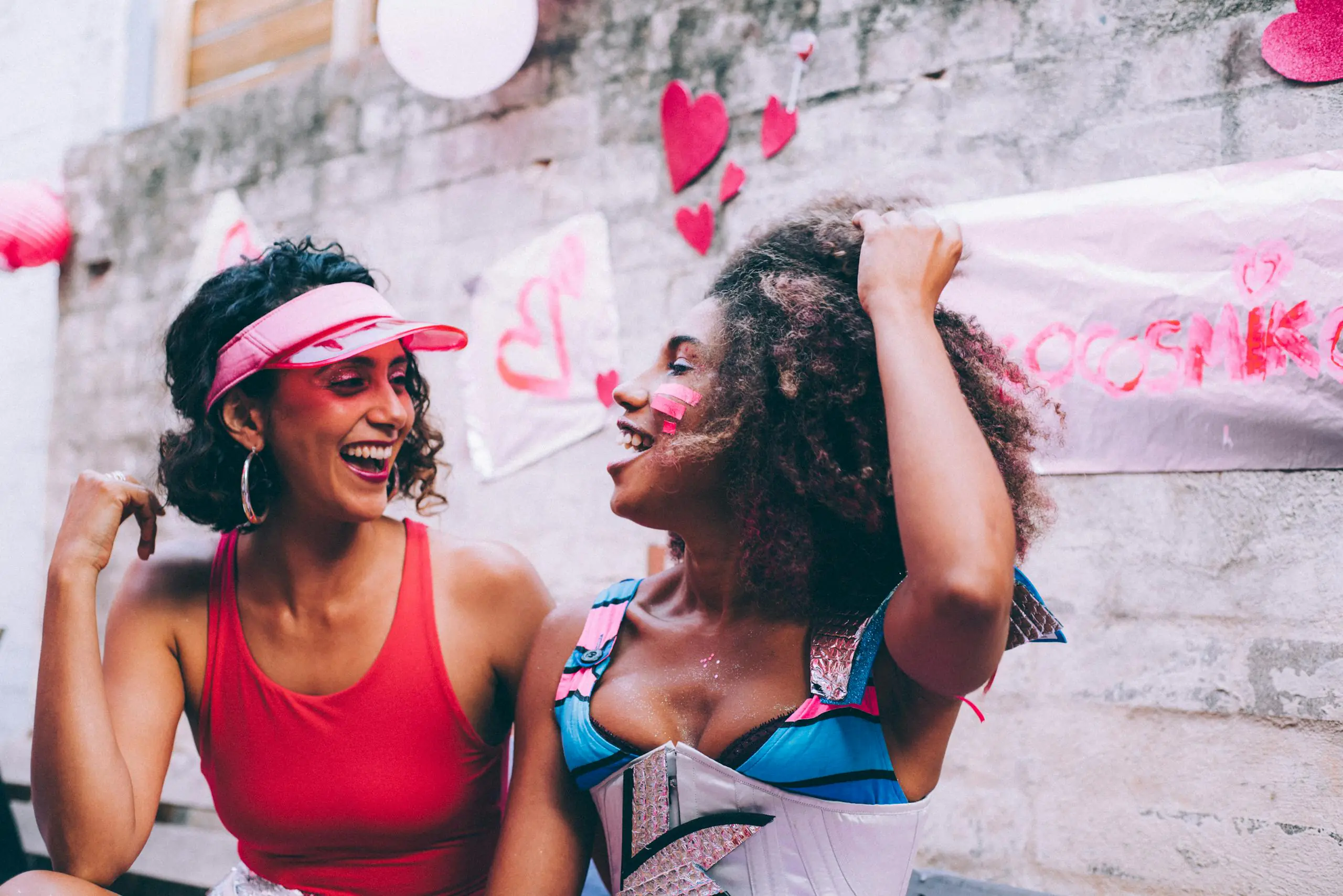 Two women laughing together at colorful outdoor celebration with heart decorations.