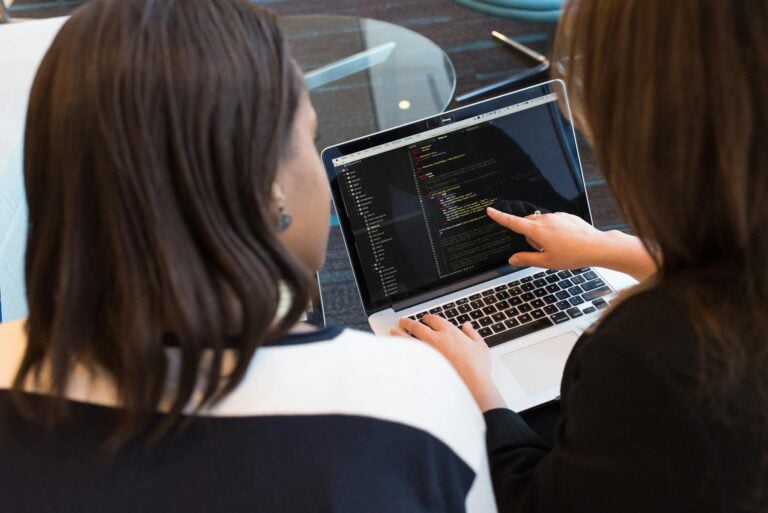 Two women collaboratively working on software programming code indoors together.