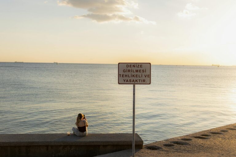 Woman sitting by the sea at sunset in Istanbul with water entry warning sign visible.