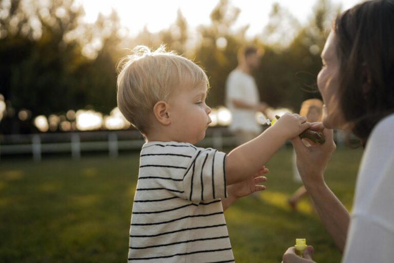Young child blowing bubbles with adult in sunny park setting, playful moment.
