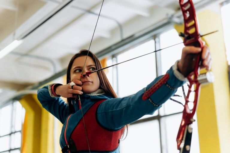 Young female archer aiming bow and arrow indoors with concentration.