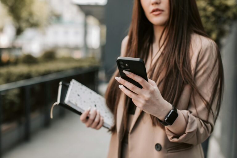 Young woman in beige blazer checking smartphone while holding notebook outside