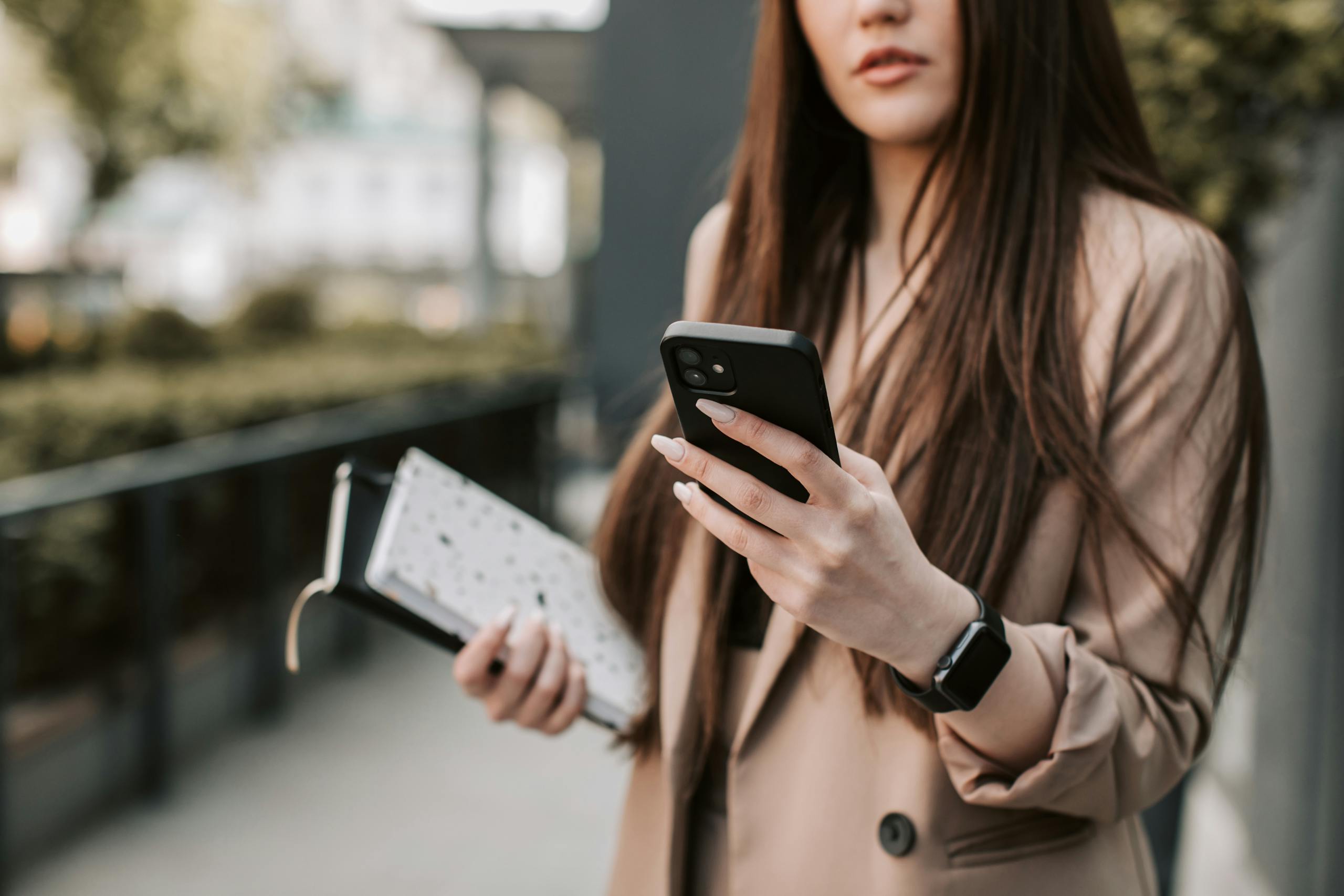 Young woman in beige blazer checking smartphone while holding notebook outside