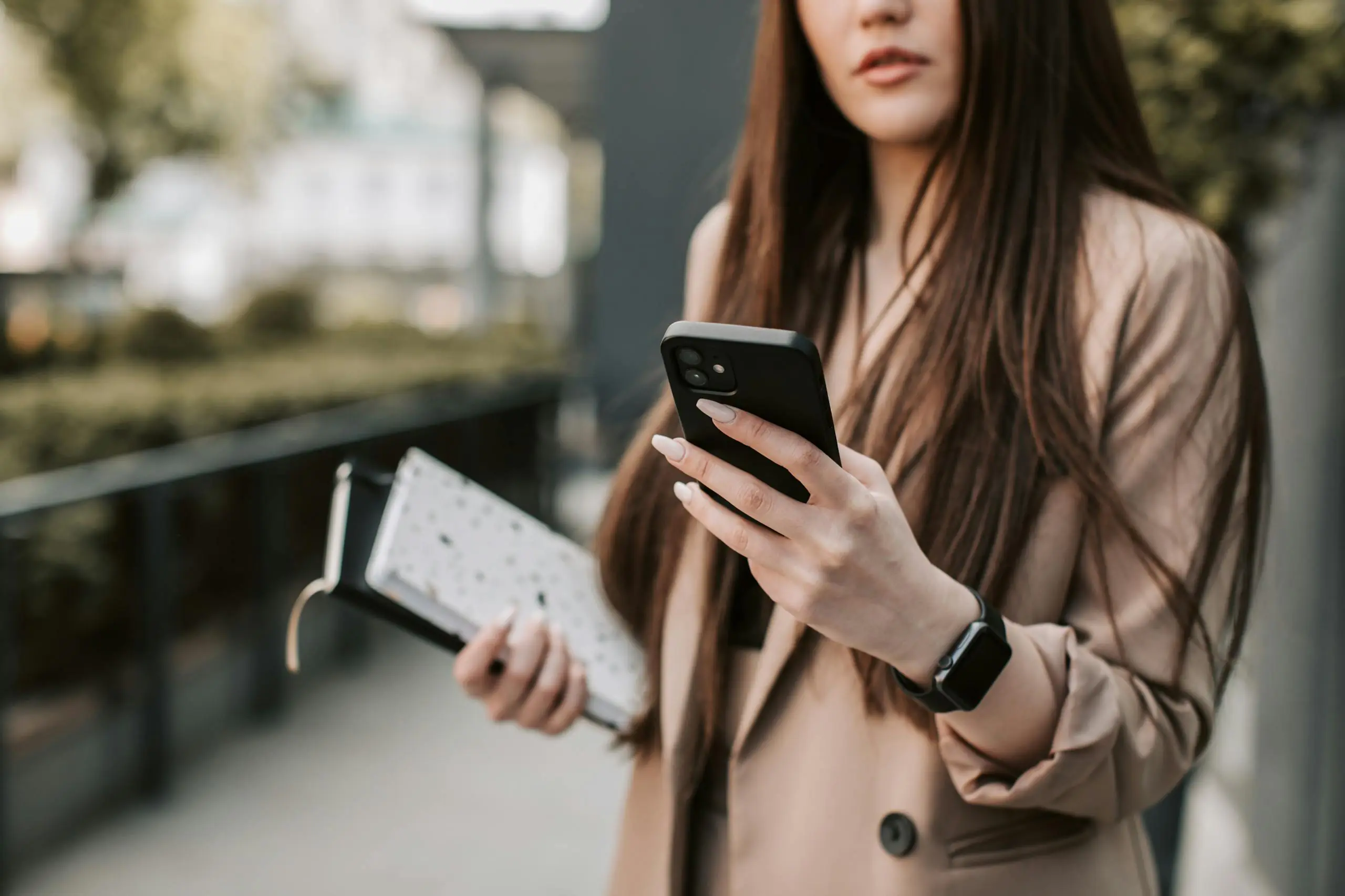 Young woman in beige blazer checking smartphone while holding notebook outside